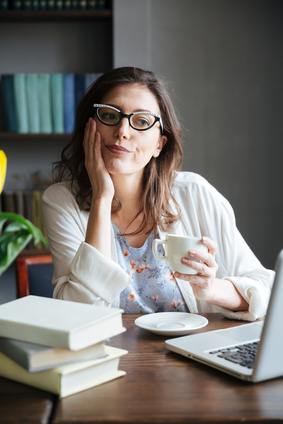 Bored woman in glasses holding cup of tea looking at laptop sitting at home